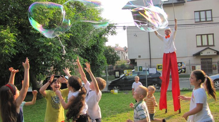 Tłumny wieczorny koncert na świeżym powietrzu, światła sceny rzucają ciepłe bursztynowe i fioletowe refleksy na wiwatującą publiczność, energetyczna atmosfera, zarys zespołu występującego pod girlandami świateł