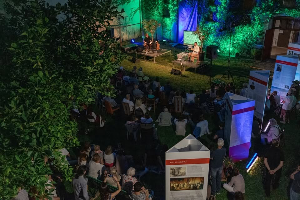Crowded evening outdoor concert with stage lights casting warm amber and purple hues over cheering festival-goers, energetic atmosphere, and silhouetted band performing under string lights