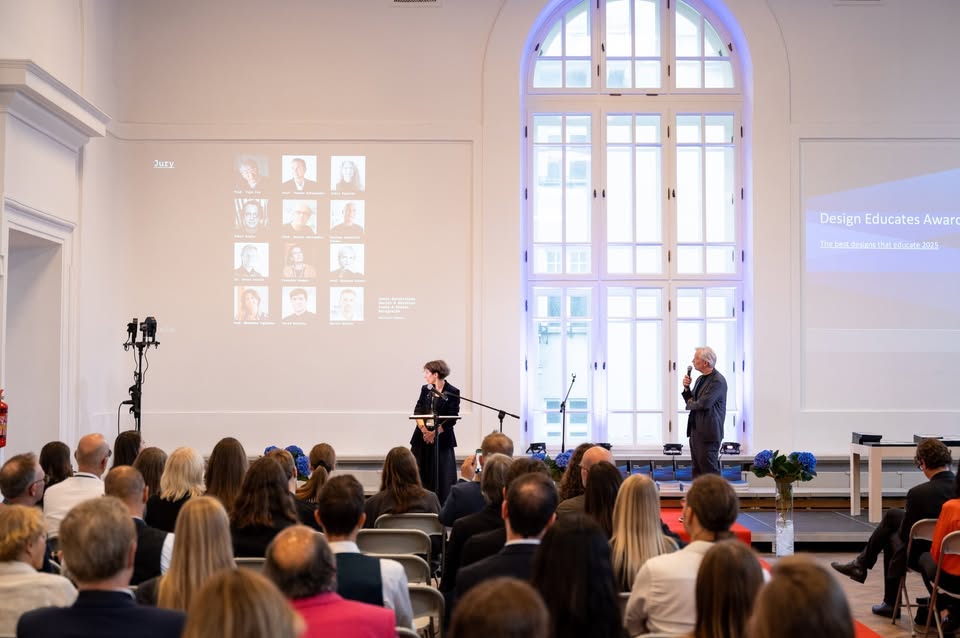 Gala Design Educates Awards w eleganckiej białej sali, dwóch prelegentów na scenie prezentujących członków jury na ekranie projekcyjnym przed siedzącą publicznością