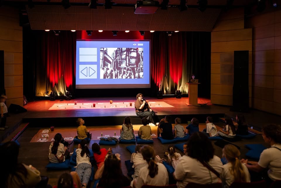 Children sitting on blue floor cushions during an interactive storytelling or educational presentation in a modern auditorium, with a presenter on stage and a graphic novel illustration displayed on a large projection screen