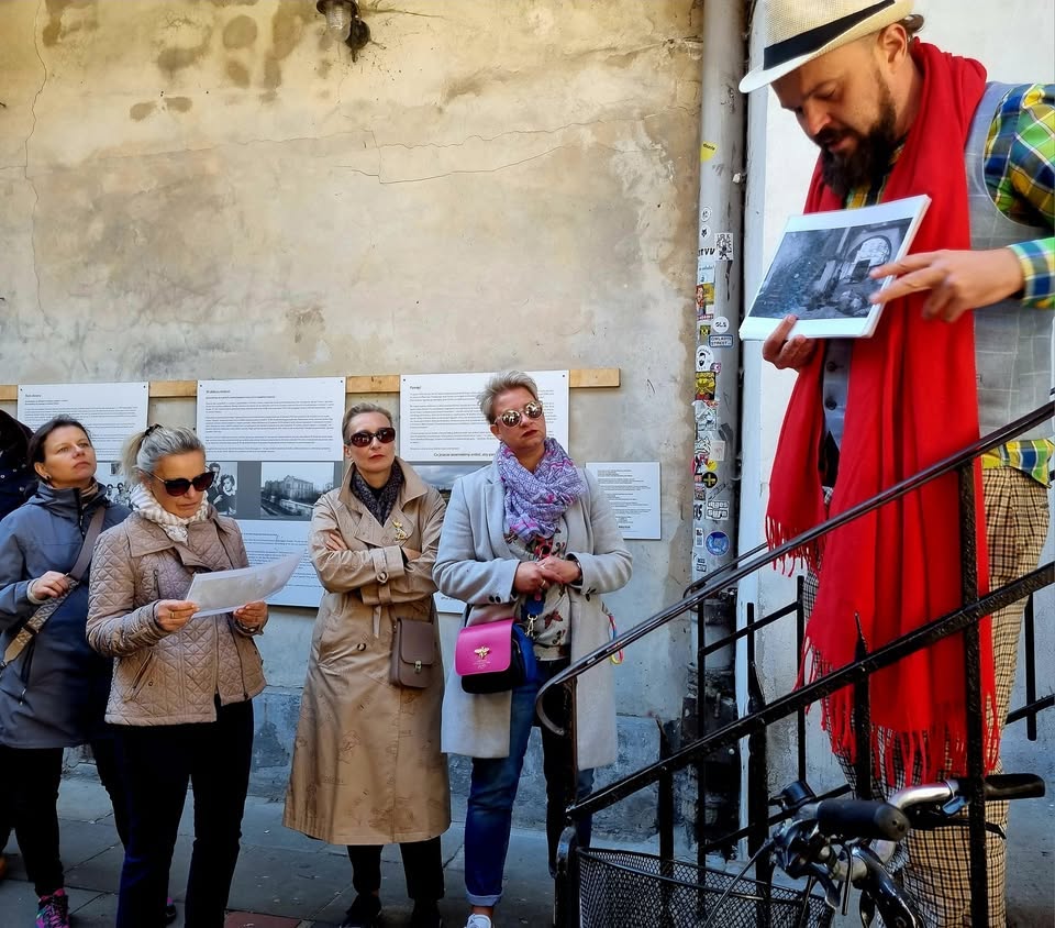 A colorfully dressed tour guide wearing a red scarf and straw hat presents a historical photograph to a group of attentive women during an outdoor city walking tour near an old building with exhibition panels